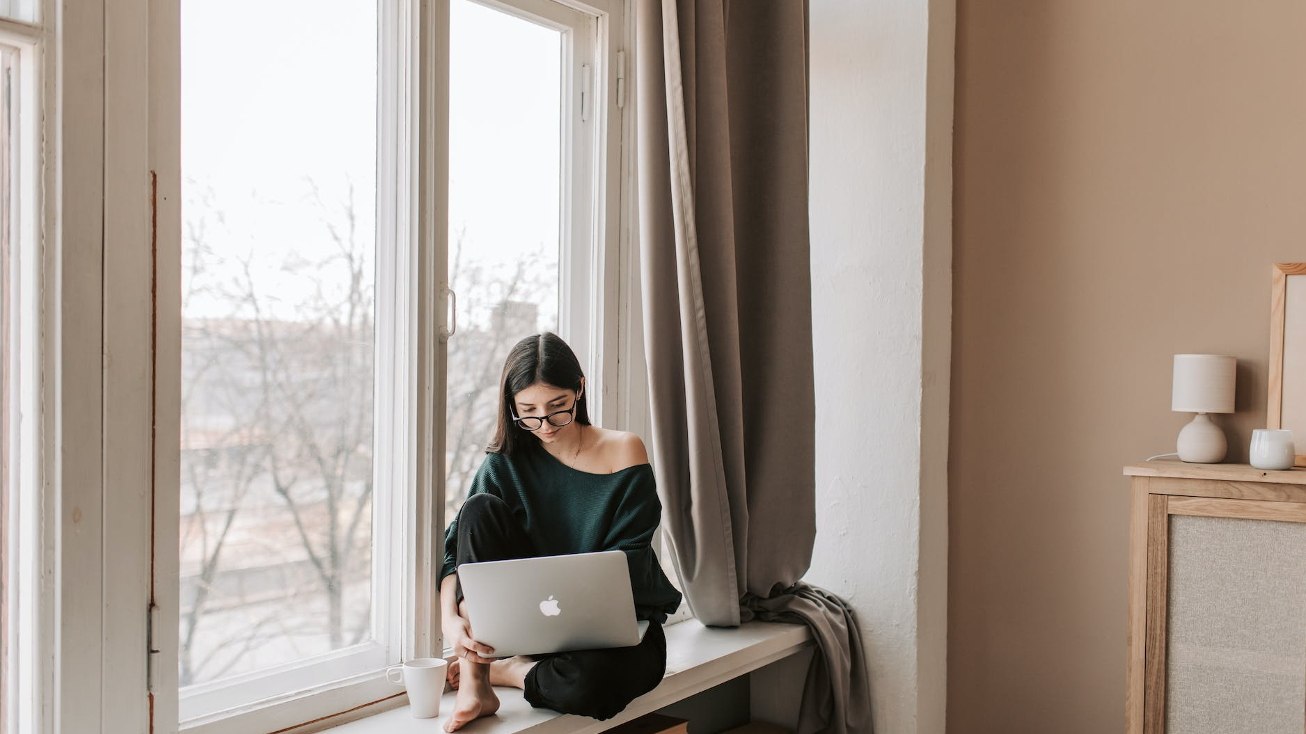 young female freelancer sitting with laptop on windowsill at home