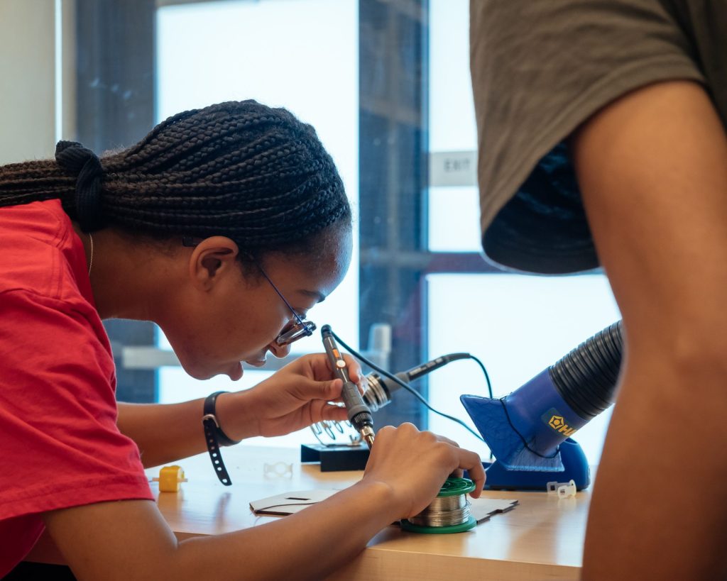 A person with braided hair is focused on soldering a component on a workbench, using a soldering iron. Tools and materials, including solder and a soldering station, are visible in the workspace.