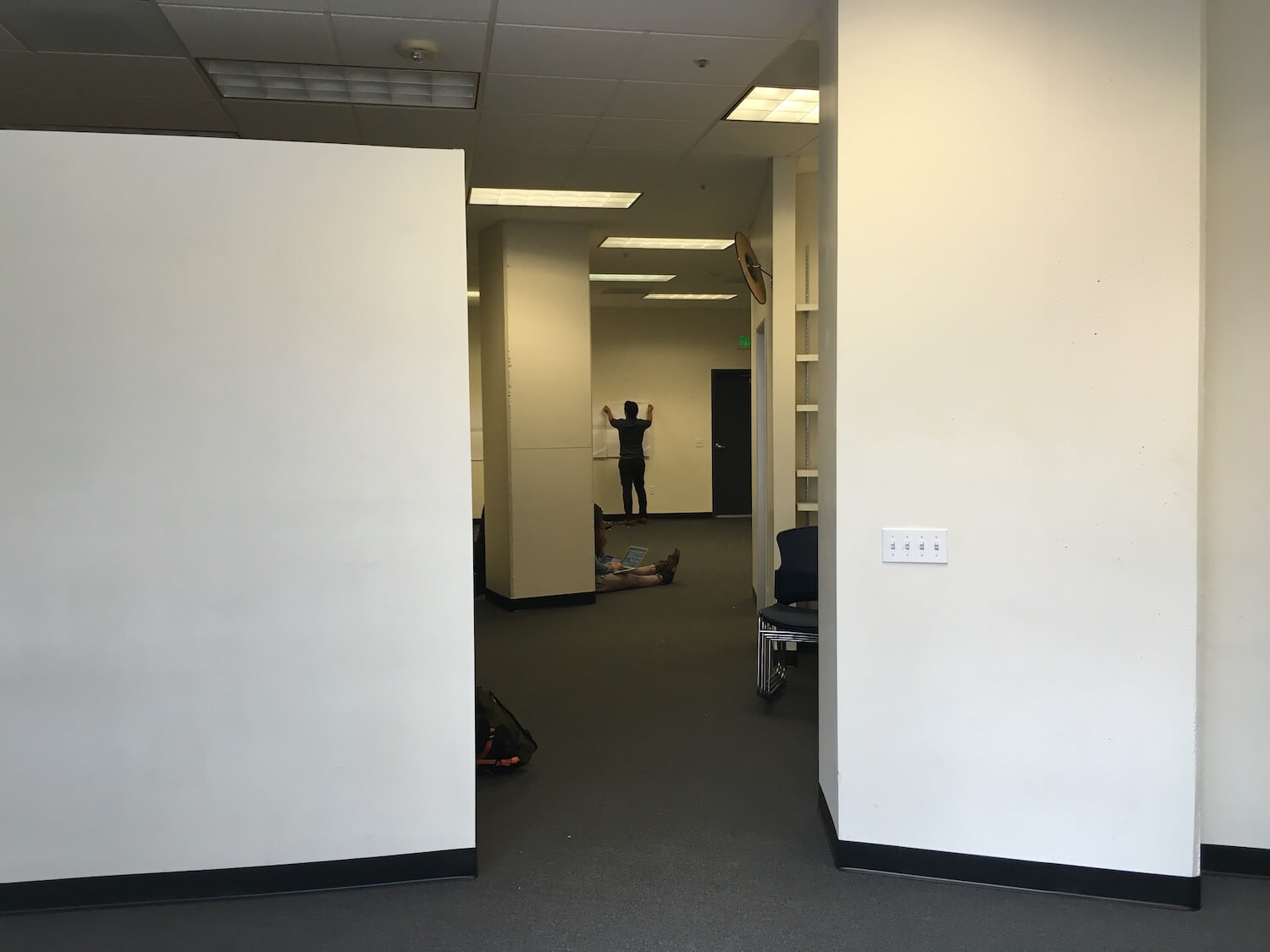An interior view of an empty office space, featuring white walls, fluorescent lighting, and a floor with dark carpeting. In the background, a person is seen standing with their arms raised, while another individual is lying on the floor.